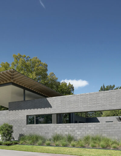 Modern single-story house with an overhanging flat roof, large windows, and concrete brick facade, set against a backdrop of trees and a clear sky.