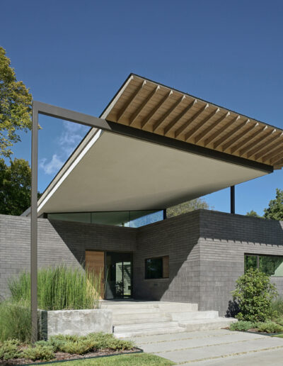 Modern house with geometric design and cantilevered roof, featuring a brick facade and large windows, surrounded by lush trees.