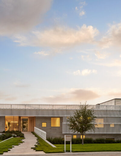 Modern two-story building with large glass windows, a sleek exterior, and a ramp leading to the entrance. Surrounded by green grass and trees, under a partly cloudy sky.